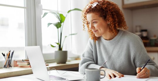 Cute university student with curly red hair doing homework sitting at kitchen table with coffee cup and wired earphones next to big window, watching online tutorial on laptop and writing in copybook