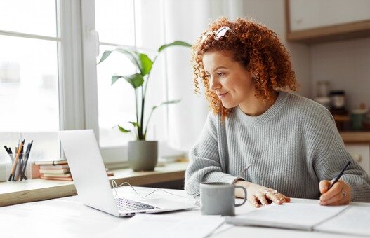 Cute university student with curly red hair doing homework sitting at kitchen table with coffee cup and wired earphones next to big window, watching online tutorial on laptop and writing in copybook