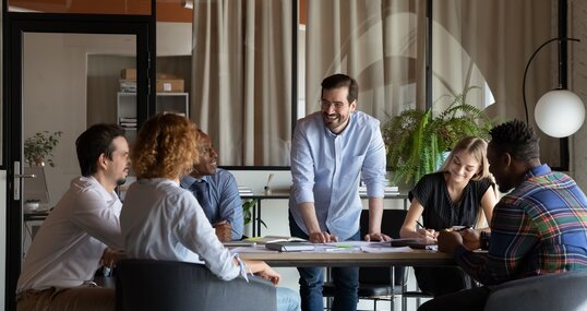 Happy male business team leader talking to employees at corporate meeting, discussing work project, sharing jokes, laughing. Positive mentor training diverse group of interns, having fun