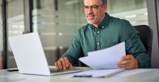 Smiling middle aged executive, mature male hr manager holding documents using laptop looking at pc computer in office at desk, checking financial data in report, doing account paper plan overview.