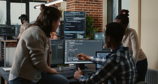 Programer sitting on desk discussing with mixed team of software developers about artificial intelligence innovation. Programmers doing teamwork looking at running machine learning algorithms.