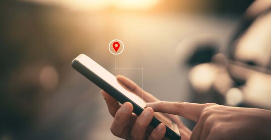 Foto einer Frauenhand die ein Smartphone benutzt mit einem Straßenhintergrund mit einer Grafik eines Navigationspunktes.  