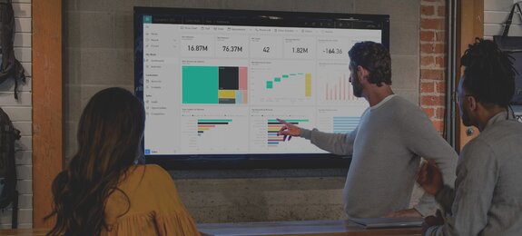 Drei Personen stehen vor einem Bildschirm und unterhalten sich über Diagramme, die dort abgebildet sind. Die Diagramme sind dabei in dem CRM-System Microsoft Dynamics 365 sichtbar.