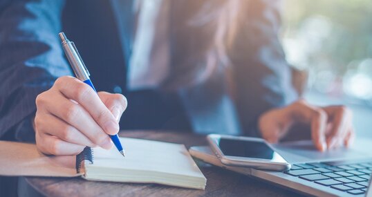 Business woman hand writing on a notepad with a pen and using laptop in the office.