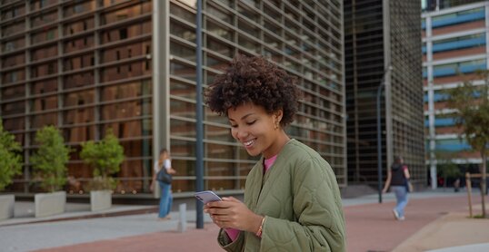 Sideways shot of beautiful woman with Afro hair holds smartphone reads received sms message has cheerful expression sits in downtown against cityscrapers dials number of taxi service. Technology