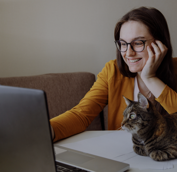 Remote work. The girl is sitting at the computer and looks at screen. A tabby cat sits next to me and also looks at screen. On a white table is a potted flower. A person sincerely smiles at someone. 