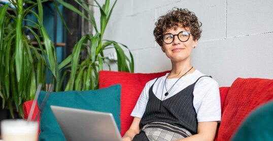 Happy young female in elegant casualwear sitting on couch with laptop in front