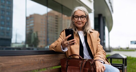 an older woman with a mobile phone sits on a bench outside