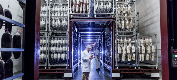 Foto einer Fabrikhalle der Bell Food Group mit zwei Mitarbeitern in Schutzkleidung und Waren die von Regalen hängen